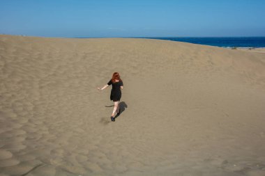 Genç bir kadın mavi gökyüzü ile denize doğru kum tepeciği koşuyor.