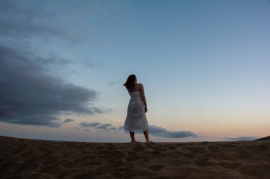 Genç bir kadın sırtı kum tepelerinde duruyor.