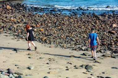 Kısa siyah elbiseli genç bir kadın ve deniz kenarında taşlı bir plajda bir çocuk.
