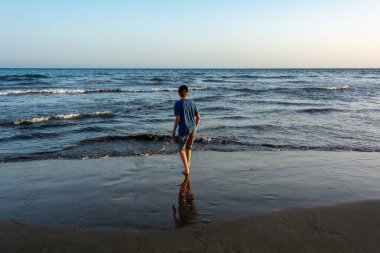 Şort ve kep giyen bir çocuk denizin kıyısındaki kumlu bir sahilde duruyor, mavi bir gökyüzü var.