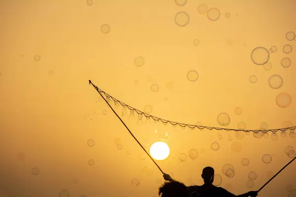 A man in silhouette makes lots of soap bubbles with a soap bubble stick, they float in the orange sky at sunset