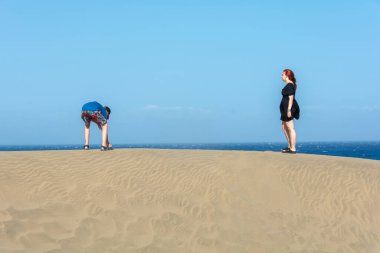 Deniz ve mavi gökyüzü ile kum tepeciğinde duran iki genç.