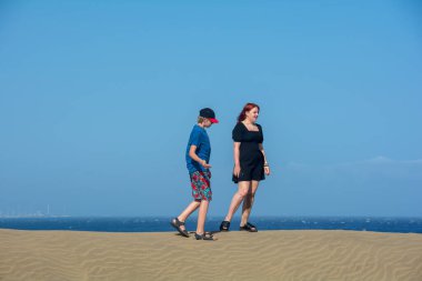 Deniz ve mavi gökyüzü ile kum tepeciğinde duran iki genç.