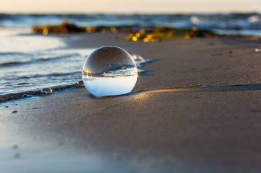 A glass ball lies in the waves on the sandy beach, the sea and the setting sun are reflected in the ball clipart