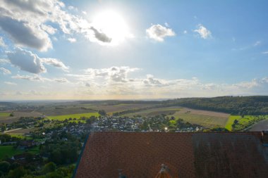 Eski bir şatonun çatısı ve içinde köy, tarım arazisi, orman ve mavi gökyüzü olan yeşil bir manzara.