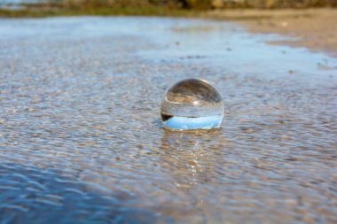 Camdan bir top kumlu sahilde dalgalarda yatıyor, deniz ve batan güneş topa yansıyor.