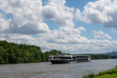 Nehir boyunca yeşil doğa, mavi gökyüzü ve beyaz bulutlarla bir eğlence gemisi yol alır.