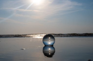 Camdan bir top kumlu sahilde dalgalarda yatıyor, deniz ve batan güneş topa yansıyor.