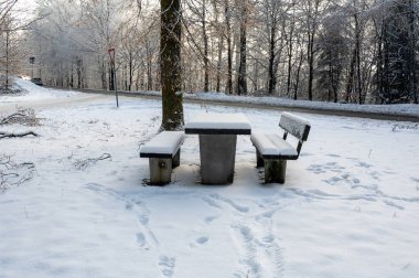 Picnic bench and table in a parking lot in the snow, in a forest  clipart