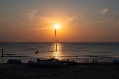 Yelkenliler kumlu sahilde uzanır, güzel turuncu günbatımıyla denizin üzerinde.