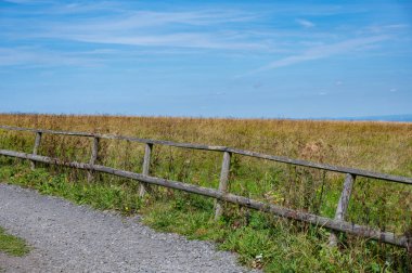 Geniş bir çayırın önündeki ahşap çitli manzara, yolu ve mavi gökyüzü.