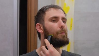The man shaves his big beard. The guy shaves off his big beard. Young guy shaves his beard at home. Personal care.