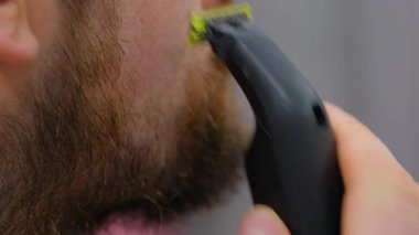 A man shaves his beard with an electric razor. The guy shaves off his big beard. Young guy shaves his beard at home. Personal care.