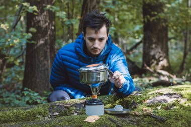 Mavi ceketli bir adam portatif bir sobada çay ve yemek için su kaynatıyor ve vahşi doğanın ortasında paslanmaz çelik bir tencerede. Yürüyüş yaşam tarzı.
