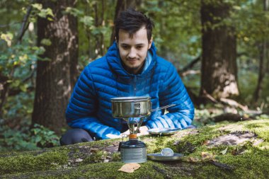 Mavi ceketli bir adam portatif bir sobada çay ve yemek için su kaynatıyor ve vahşi doğanın ortasında paslanmaz çelik bir tencerede. Yürüyüş yaşam tarzı.