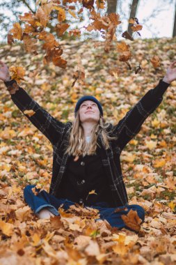 Gülümseyen esmer, meşe ormanındaki renkli yaprakların arasında oturmuş yaprak fırlatıyor. Sonbahar mevsiminde sonbahar kıyafetleri içinde gerçek bir kadının samimi portresi..