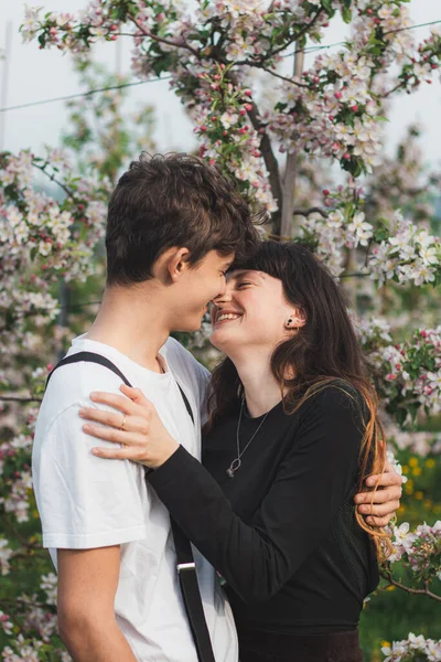 リンゴの木の下を歩く2人の若者の間の美しい強い愛 カジュアルな服を着たカップルの肖像画を描いた 若いカップルの笑顔 — ストック写真