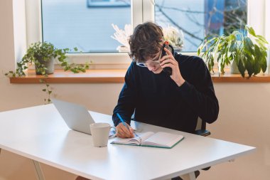 goal-oriented brown-haired businessman is on the phone with his supervisor, jotting notes in his diary. Making and arranging an appointment with a client. A young visionary in his office.