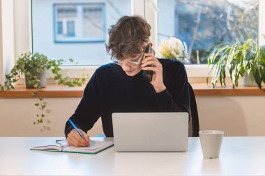 Purposeful brown-haired businessman is on the phone with an exiting client, jotting notes in his diary. Online business meeting with client. Young visionary.
