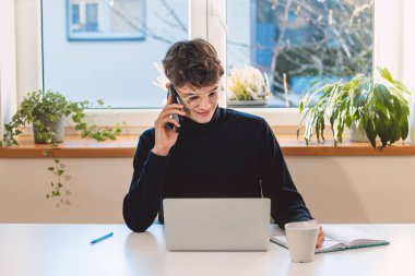 Purposeful brown-haired businessman is on the phone with an exiting client, jotting notes in his diary. Online business meeting with client. Young visionary.