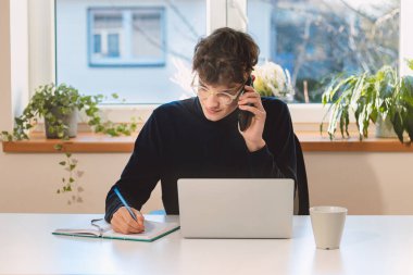 Purposeful brown-haired businessman is on the phone with an exiting client, jotting notes in his diary. Online business meeting with client. Young visionary.
