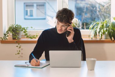 Purposeful brown-haired businessman is on the phone with an exiting client, jotting notes in his diary. Online business meeting with client. Young visionary.