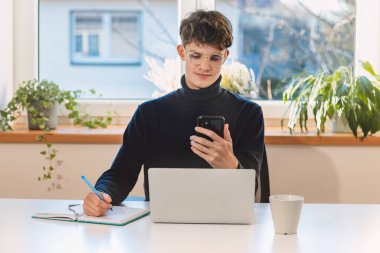 Purposeful brown-haired businessman is working on his laptop and jotting down his notes from the interview on his cell phone. The businessman is performing daily tasks.