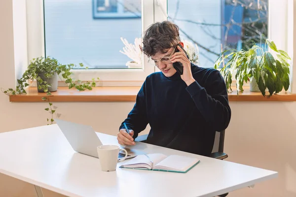 Purposeful brown-haired businessman is on the phone with an exiting client, jotting notes in his diary. Online business meeting with client. Young visionary.