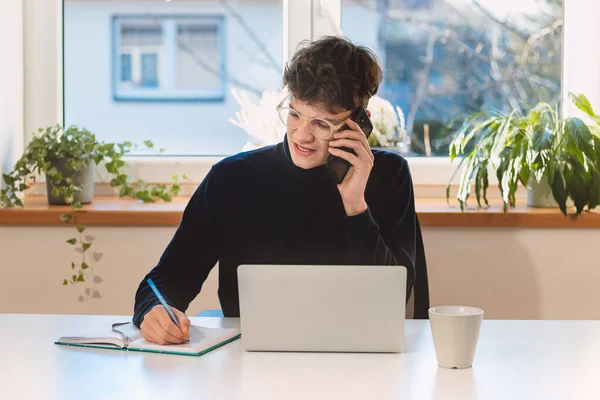 Purposeful brown-haired businessman is on the phone with an exiting client, jotting notes in his diary. Online business meeting with client. Young visionary.