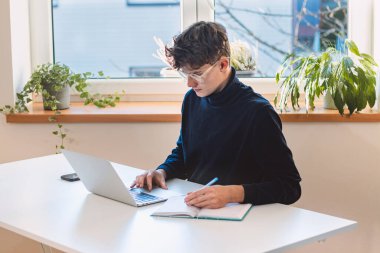 Entrepreneur in his study by the window is thinking of a new concept and strategy that will lead his company to success. A young, goal-oriented guy with glasses and brown hair.