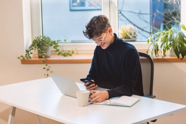 Purposeful brown-haired entrepreneur with a smile on his face answers his clients on his mobile phone in his office with ample daylight. Working from home.