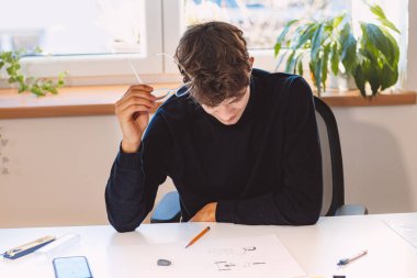 Brown-haired architect is thinking hard about a new design for a new prototype, which he draws in pencil on paper. Inventing a new design. An ambitious young boy. Illuminated workroom.