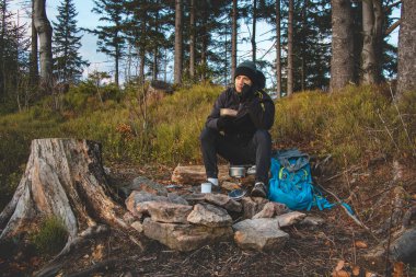 Outdoor wilderness trekking enthusiast sitting by a campfire enjoying breakfast at sunrise. Breakfast and catching vitamin D at the top of the hill. The life of a nature-loving boy.