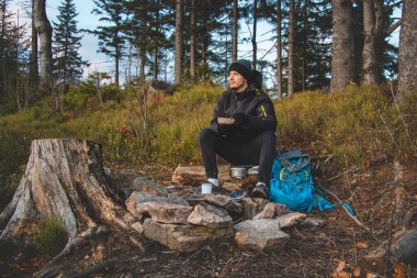 Outdoor wilderness trekking enthusiast sitting by a campfire enjoying breakfast at sunrise. Breakfast and catching vitamin D at the top of the hill. The life of a nature-loving boy.