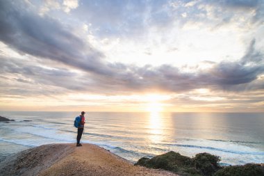 Gezgin, Portekiz 'in Algarve bölgesindeki Ophixe kasabasının yakınındaki bir uçurumun kenarında duruyor ve berrak mavi okyanusta güneşin batışını izliyor. Gökyüzündeki renklerin oyunu.