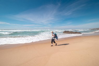 Sırt çantalı adam Praia do Almograve boyunca yüzünde bir gülümsemeyle yürüyor. Taşınmanın ve yeni yerler keşfetmenin keyfi. Odemira bölgesi, batı Portekiz. Balıkçı Patikası boyunca gezinen, Rota Vicentina.
