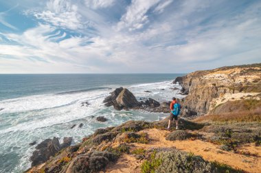 Portekiz 'in batısındaki Odemira bölgesinde Atlantik Okyanusu' na bakan bir uçurumun kenarında duran genç bir sırt çantasının neşeli ifadesi. Balıkçı Patikası boyunca gezinen, Rota Vicentina.