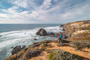 Portekiz 'in batısındaki Odemira bölgesinde Atlantik Okyanusu' na bakan bir uçurumun kenarında duran genç bir sırt çantasının neşeli ifadesi. Balıkçı Patikası boyunca gezinen, Rota Vicentina.