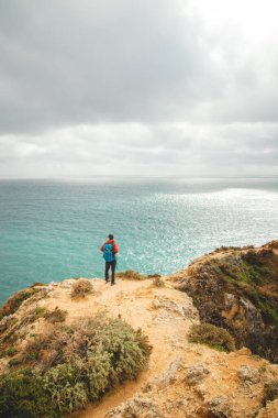 Sırt çantalı maceraperest bir uçurumun kenarında duruyor ve Güney Portekiz 'in Algarve bölgesindeki Lagos' taki Punta de la Piedad 'ın güzel sarı-altın kayalık grubunu gözlemliyor..