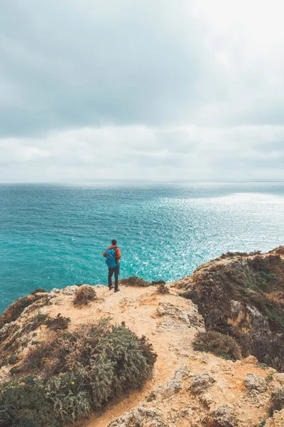 stock image Adventurer with a backpack stands on the edge of a cliff and observes the beautiful group of yellow-gold rocks of Punta de la Piedad in Lagos, in the Algarve region of southern Portugal.