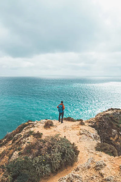 stock image Adventurer with a backpack stands on the edge of a cliff and observes the beautiful group of yellow-gold rocks of Punta de la Piedad in Lagos, in the Algarve region of southern Portugal.