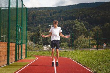 Beyaz tişört ve siyah şort giyen kahverengi saçlı bir çocuk atletik bir ovalda ip atlıyor. Atlama, koordinasyon ve dayanıklılık gücünü geliştirmek için eğitim.