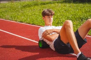 Atletik oval vücut yapısı olan kahverengi saçlı çocuk daha iyi iyileşmek için kaslarına köpük silindirle masaj yapıyor. Egzersiz sonrası.