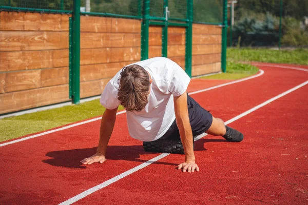 Stock image Brown-haired boy with an athletic physique on an athletic oval massages his muscles with a foam roller for better recovery. Post-workout.