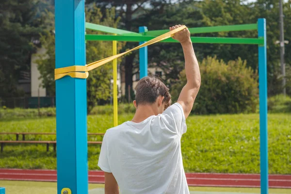 Vücudu üzerinde direnç lastiği kullanarak çalışan hedef odaklı genç, dayanıklılık eğitimi alıyor. Spor sezonuna hazırlanıyorum. Kendi vücudunla egzersiz yapıyorsun. Açık hava antrenman parkı. Bileşenler.