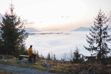 Ahşap bir bankta oturan maceraperest, altında yuvarlanan bulutları ve turuncu-sarı gün batımlarını izliyor. Adam düşünür ve meditasyon yapar. Beskydy Dağları.