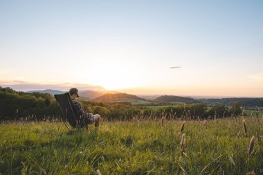Ekose gömlekli genç adam seyyar bir hafif koltukta oturur, vadinin manzarasının ve ufukta batan güneşin tadını çıkarır. Beskydy Dağları, Çek Cumhuriyeti.
