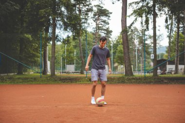 20 'li yaşlardaki futbolcu kil sahada bir balonla oynuyor. Amatör spor. Bireysel futbol eğitimi. - Futbol. Tekme, vuruş, pas ve top sürme.