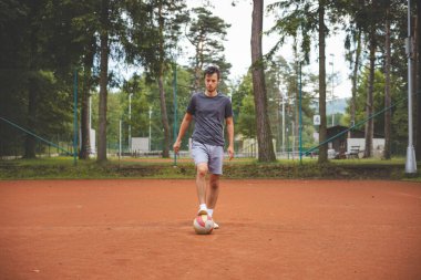 20 'li yaşlardaki futbolcu kil sahada bir balonla oynuyor. Amatör spor. Bireysel futbol eğitimi. - Futbol. Tekme, vuruş, pas ve top sürme.