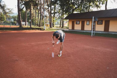 Yüzünde gülümseme olan siyah saçlı bir sporcu kil sahada badminton oynar. Amatör spora olan tutkum. Yeni hareket becerileri öğreniliyor.